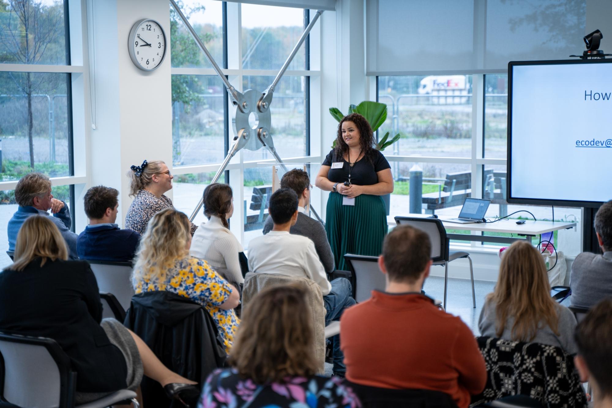 wide shot of presentation being held by a female person to meeting room of people using a monitor to show information